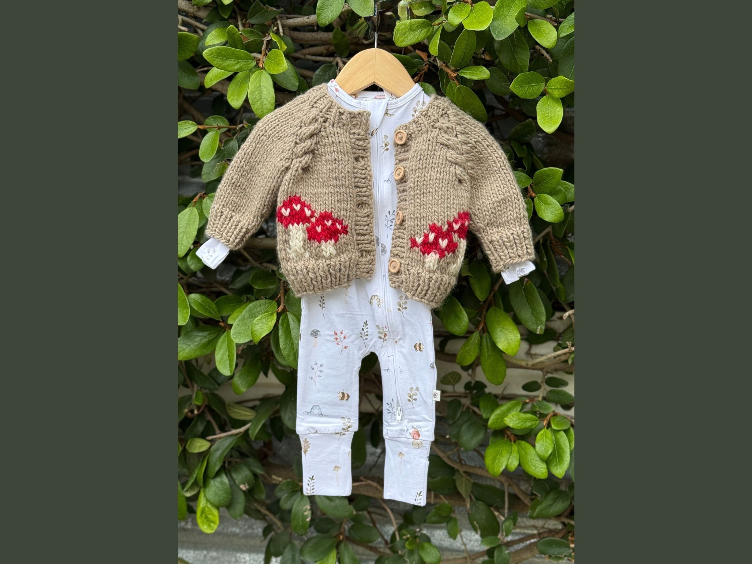 Children's knitted cardigan with floral patterns and white pants hanging against a green leafy background